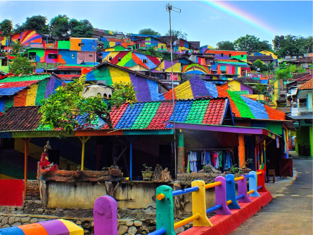 Un paesino di montagna con case dipinte con colori vivaci e motivi vari, tra cui blu, giallo, rosso e verde, si stagliava sotto un cielo limpido su cui si stagliava un arcobaleno. Una staccionata colorata costeggiava la strada.