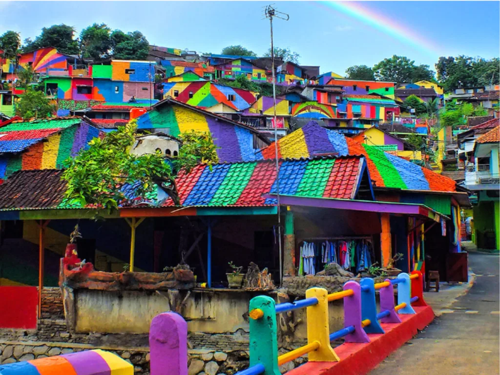 Un paesino di montagna con case dipinte con colori vivaci e motivi vari, tra cui blu, giallo, rosso e verde, si stagliava sotto un cielo limpido su cui si stagliava un arcobaleno. Una staccionata colorata costeggiava la strada.